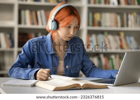 Serious hipster red haired student girl in big headphones studying in college library, writing notes at laptop, open book, listening to music for focus, watching learning class, webinar, lesson Royalty-Free Stock Photo #2326376815