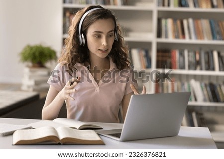 Focused pretty student girl in headphones talking on video call at laptop in university library, sitting at computer, open books, consulting online teacher, attending learning Internet conference Royalty-Free Stock Photo #2326376821