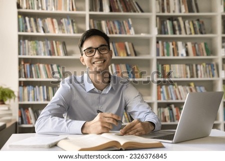 Cheerful nerdy student guy in eye glasses looking at camera, smiling, laughing, getting knowledge, university higher education, posing at table with open book, laptop in campus library Royalty-Free Stock Photo #2326376845