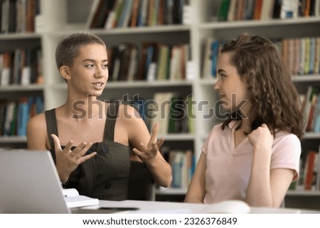 Two positive young school student girls cooperating on home assignment in college campus together, talking at laptop in library, discussing creative ideas for research study Royalty-Free Stock Photo #2326376849