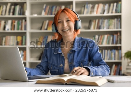 Cheerful hipster student girl with teeth brackets wearing headphones, looking at camera, smiling, working in university library, writing essay, article on laptop computer. Posing for portrait