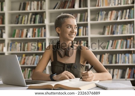Positive young hipster student girl doing academic research study in university library, working on home assignment, article, essay, writing notes at textbook, laptop, looking away, thinking, smiling Royalty-Free Stock Photo #2326376889