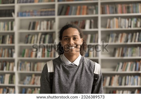 Cheerful long haired fresh African American student guy with backpack standing in campus, university library with bookshelves in background, looking at camera with toothy smile. Head shot portrait
