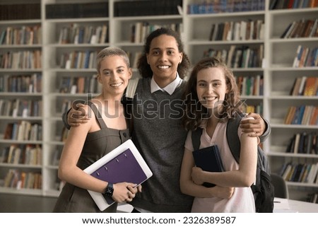 Happy diverse high school friends enjoying meeting in campus library, posing with books on shelves in background, hugging, looking at camera, smiling, enjoying friendship, education
