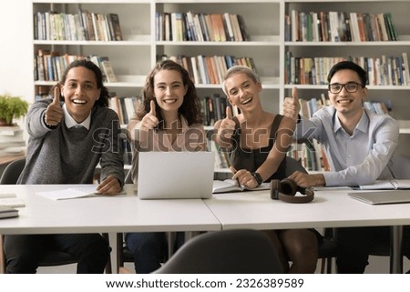 Cheerful divers young university students posing in campus library, sitting at table with bookshelves in background, looking at camera, showing like thumb up hands, laughing, enjoying education
