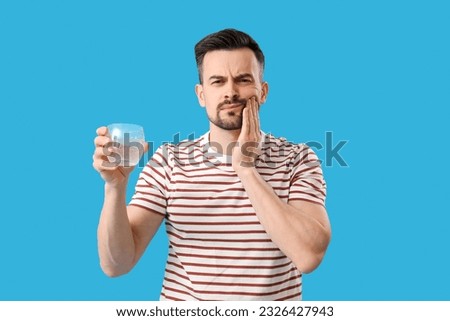 Young man with dissolved tablet in glass of water suffering from toothache on blue background