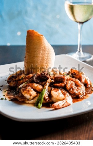 Blackened shrimp tossed in red pepper sauce and fresh chives on bed of parmesan grits with fresh baguette on white plate with cold white wine glass in the blue background. Royalty-Free Stock Photo #2326435433
