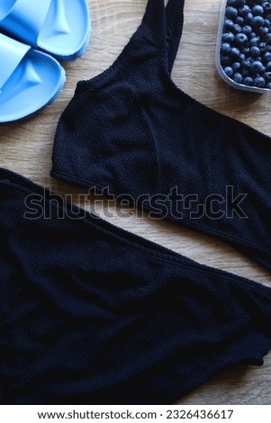 Black swimsuit, striped towel, straw hat, bowl of blueberries and blue sandals. Summer or beach essentials. Top view, wooden background.