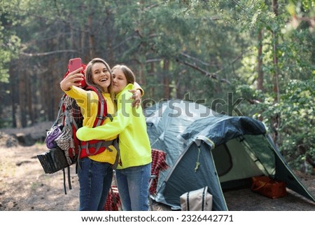 Mom with daughter taking a selfie in a forest camp. They standing near the tent and take a photo wit smartphone. Motherhood and trust of the younger generation.