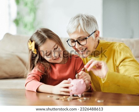 Grandmother and her granddaughter Putting Coin Money In Piggybank  At Home. Personal Savings, Bank Safety And Financial Investments Concept Royalty-Free Stock Photo #2326488613