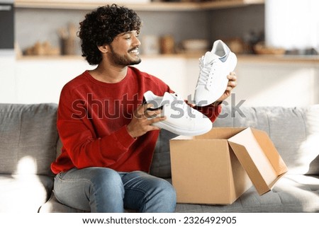 Portrait of happy young indian man unboxing parcel with shoes at home, smiling eastern guy opening cardboard box and looking at new pair of white sneakers, satisfied with online shopping Royalty-Free Stock Photo #2326492905