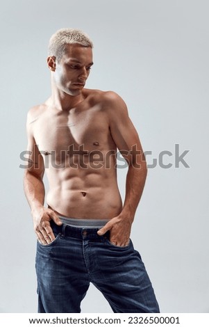 Portrait of handsome young man with blonde hair, musuclar fit body posing shirtless in jeans against grey studio background. Concept of male natural beauty, body care, health, sport, fashion, ad Royalty-Free Stock Photo #2326500001