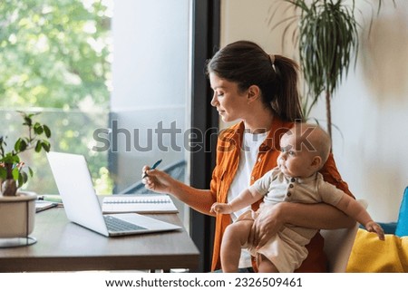 Female entrepreneur writing in notes while working on laptop and taking care of her baby at home. Royalty-Free Stock Photo #2326509461