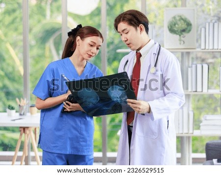 Asian handsome professional successful male doctor in white lab coat with stethoscope holding xray film talking brainstorming with female assistant nurse in blue uniform in clinic hospital office.