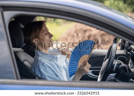 Tired exhausted middle aged woman waving blue fan suffers from stuffiness driving car on summer hot weather. Overheating, sultriness, high temperature, swelter in car with broken air conditioner. Royalty-Free Stock Photo #2326542915
