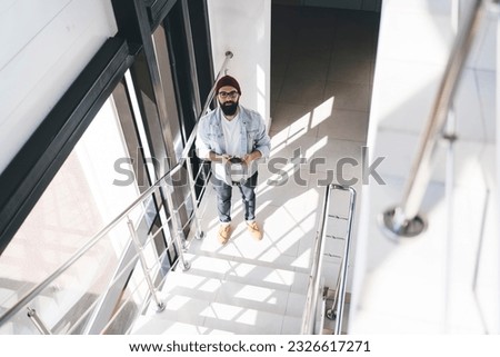 From above full body of young hipster in casual clothes hat and glasses browsing smartphone while having free time standing near staircase looking at camera