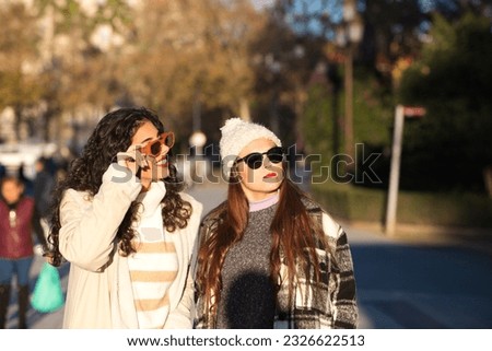 two young, beautiful, Spanish women who are friends dressed in winter clothes and wearing sunglasses are walking and enjoying the sunny day in seville, spain.