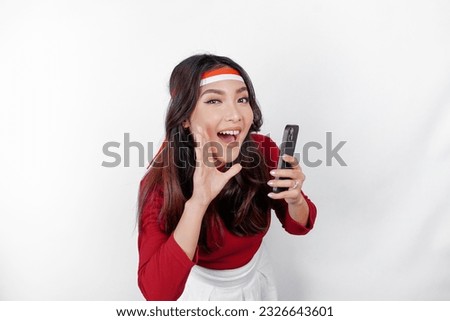 Young beautiful woman wearing a red top is holding her phone while shouting and screaming loud with a hand on her mouth. Indonesia's independence day concept. Royalty-Free Stock Photo #2326643601