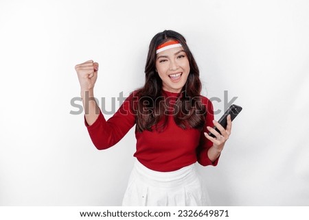 A young Asian woman with a happy successful expression while holding her phone and wearing red top and flag headband isolated by white background. Indonesia's independence day concept.