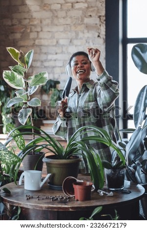 Morning routine and mediation, mental health concept. Cozy green interior. Attractive plus size African American young woman taking care about home plants. Enjoying household activity, singing, dance