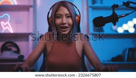 Portrait Young Asian woman professional gamer wearing headphone to gaming on pc and talking with team on microphone with fun while competition, cast gameplay or record a podcast in neon light room