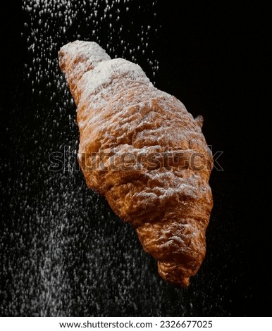 Powdering and snowing sugar to a croissant pictured in a photo studio with dark background.