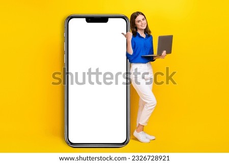 Full length photo of pretty shiny girl dressed blue shirt pointing thumb back modern device empty space isolated yellow color background