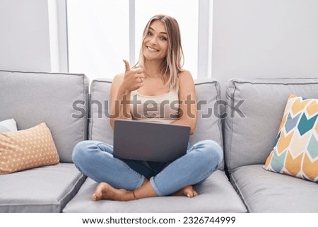 Blonde caucasian woman using laptop at home sitting on the sofa smiling happy and positive, thumb up doing excellent and approval sign 