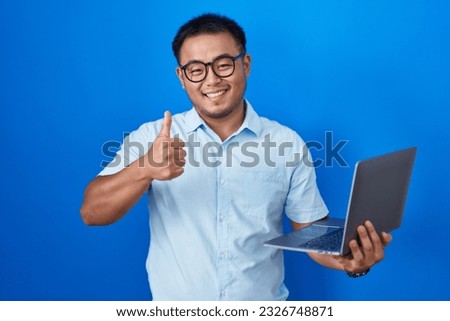 Chinese young man using computer laptop doing happy thumbs up gesture with hand. approving expression looking at the camera showing success. 