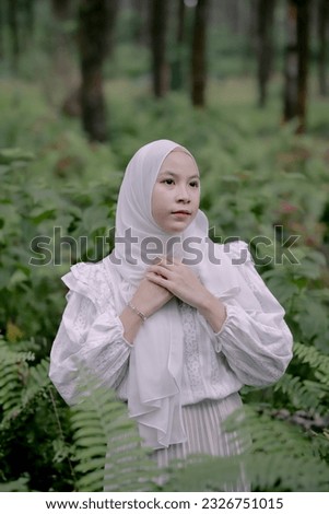 A Muslim woman dressed in white and wearing a hijab is praying in the middle of a forest. With a hopeful face.