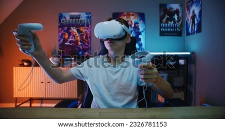 Young gamer in VR headset plays virtual reality online video game using wireless controllers. Boy having fun in leisure time in stylish neon room. Gaming at home. View from PC screen perspective. Royalty-Free Stock Photo #2326781153