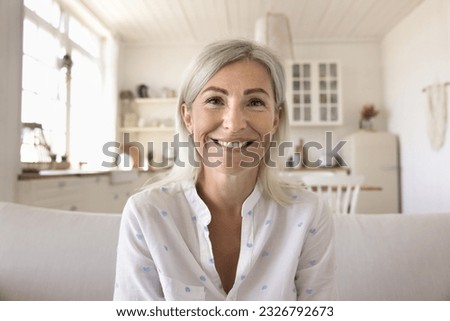 Positive pretty blonde mature woman home head shot portrait. Happy senior lady talking on online video conference call, looking at camera with toothy smile, sitting on couch with kitchen in background