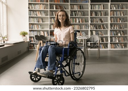 Cheerful young college student with disability, visiting campus library, holding book, looking at camera with toothy smile, promoting inclusive educational environment