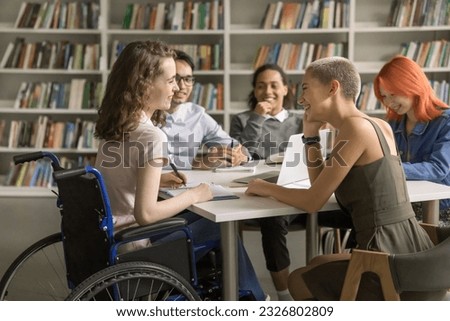 Young student girl with disability cooperating on class project in library with college friends, using wheelchair, sitting at table, talking, chatting with classmates, laughing Royalty-Free Stock Photo #2326802809
