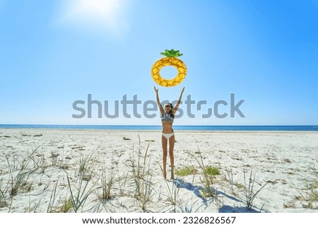 Summer bikini girl joyfully tossing an inflatable pineapple float ring against coastal backdrop. The vibrant colors, playful atmosphere, and idyllic beach Royalty-Free Stock Photo #2326826567