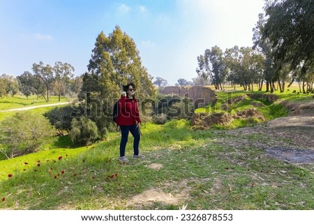 Elderly woman taking pictures of beautiful nature. Early spring in Israel. Kibbutz Beeri. Green carpet of spring grass, colorful wild flowers and swollen buds on trees. The forest plantations. 