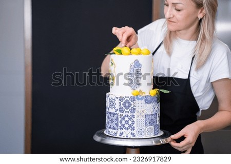 Focused housewife decorating large two-tiered cake with confit yellow lemons and green leaves cakes standing in kitchen blonde woman preparing custom confectionery for birthday party Royalty-Free Stock Photo #2326917829