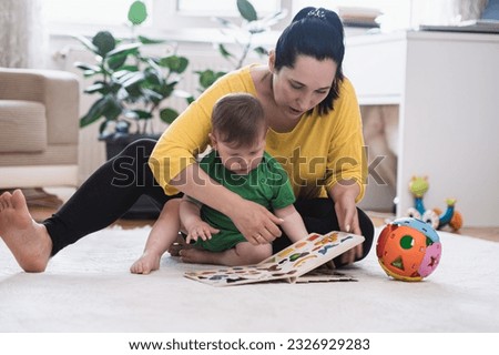 Focused mother showing little son interesting book with colorful pictures of animals sitting on white rug in living room cute baby boy developing new skills at home