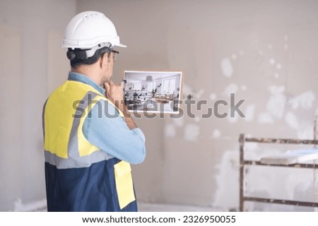 Pensive architect in white helmet looking at picture in hands stands against shabby wall in living room project design with modern Scandinavian style interior