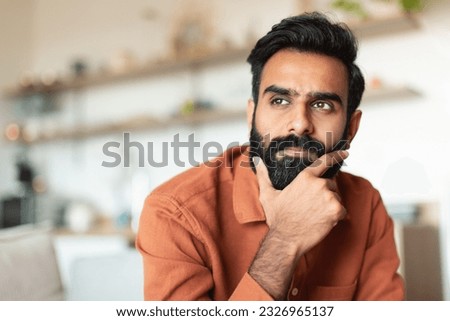 Portrait Of Pensive Young Arabic Man Thinking Sitting In Living Room At Home, Looking Aside. Thoughtful Middle Eastern Guy Touching Beard And Pondering About Issues, Free Space Royalty-Free Stock Photo #2326965137