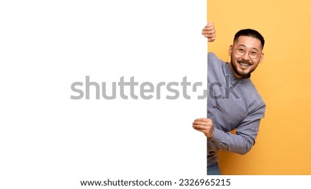 Joyful Young Asian Man Standing Behind Big White Advertisement Board, Happy Smiling Millennial Guy Demonstrating Free Copy Space On Blank Billboard, Posing Over Yellow Studio Background Royalty-Free Stock Photo #2326965215