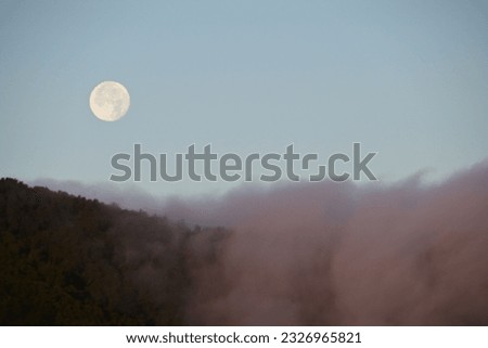 full moon cresting over costal mountain ridge with fog rolling over the top on a summer morning