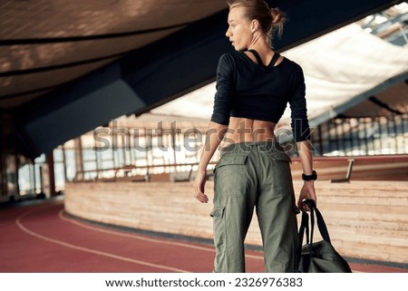 Cute young athlete girl getting ready for a workout at the stadium. Attractive slim blonde in a black top and khaki cargo pants with a sports bag goes to the locker room. Active lifestyle and sports. Royalty-Free Stock Photo #2326976383