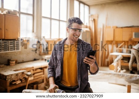 Young carpenter using a smart phone while working in his woodworking workshop Royalty-Free Stock Photo #2327027909
