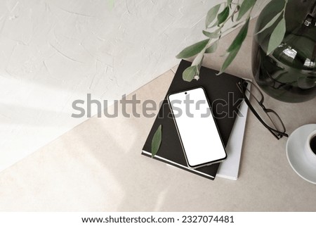 Mobile phone with empty screen mockup, notebook, glasses, vase with foliage branch, empty neutral beige background with aesthetic sunlight shadow. Minimalist business brand, social media blog template Royalty-Free Stock Photo #2327074481