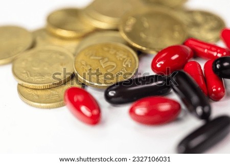 A close-up photo of Indian 5 rupees coins and red and black pills on a white backdrop. The coins are arranged in a circular pattern, with the pills placed in the center Royalty-Free Stock Photo #2327106031