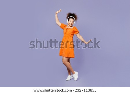 Full body young fun latin woman wear orange blouse casual clothes headphones listen to music dance raise up hands isolated on plain pastel light purple background studio portrait. Lifestyle concept