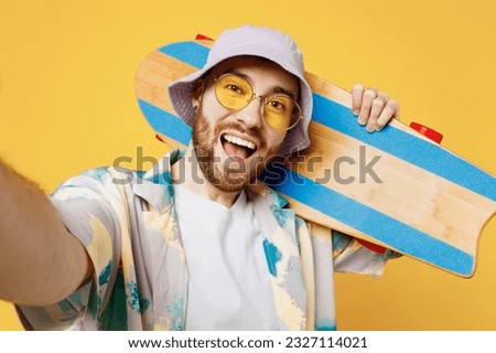 Close up young man he wear blue shirt white t-shirt casual clothes doing selfie shot pov on mobile cell phone hold inahnd skateboard pennyboard isolated on plain yellow background. Lifestyle concept
