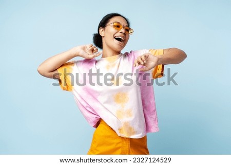 Happy African American woman dancing wearing trendy t shirt and eyeglasses isolated on blue background. Smiling stylish fashion model having fun posing for picture in studio