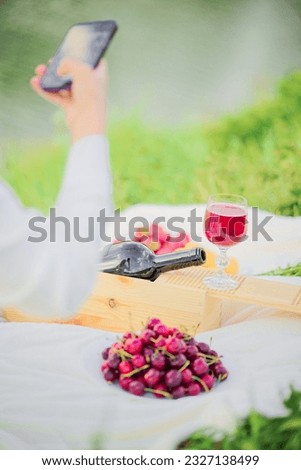 Woman taking pictures of food, wine, cherries, bottle, fruit on her smartphone at a picnic.
